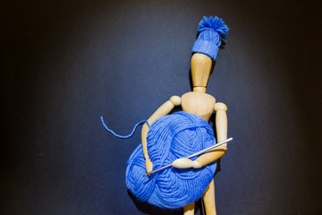 Wooden puppet doll holds a ball of blue yarn and crochet hook in its hands, and wears a knitted...