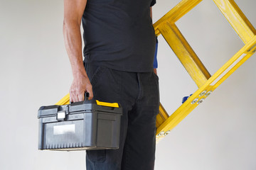Electrician holds tool box and yellow wooden ladder. The builder carries a wooden ladder and toolbox on a gray background. Breeder with tool box and yellow wooden ladder.