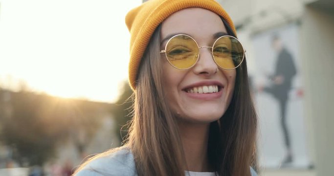 Side view on the young Caucasian girl in the sunglasses and yellow hat, turning her head, looking first serious and then smiling to the camera. Close up. Portrait. Outdoor.