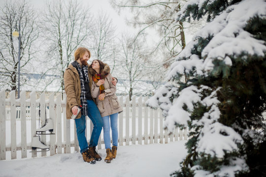 Young loving couple caucasian man with blond long hair and beard, beautiful woman have fun drinking a hot drink from thermos, eating green apple in snow park near white fence and coniferous tree