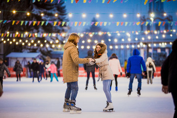 Young couple in love Caucasian man with blond hair with long hair and beard and beautiful woman have fun, active date skating on ice scene in town square in winter on Christmas Eve