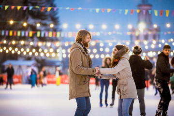 Young couple in love Caucasian man with blond hair with long hair and beard and beautiful woman have fun, active date skating on ice scene in town square in winter on Christmas Eve