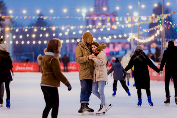 Naklejka premium Young couple in love Caucasian man with blond hair with long hair and beard and beautiful woman have fun, active date skating on ice scene in town square in winter on Christmas Eve