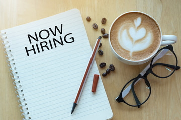 Concept NOW HIRING message on notebook with glasses, pencil and coffee cup on wooden table.