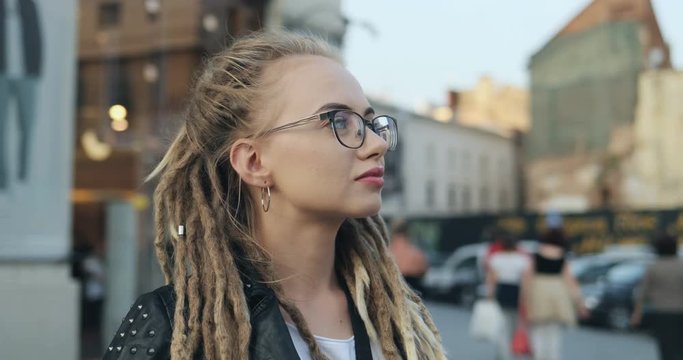 Camera zooming in on the face of a stylish Caucasian pretty girl in glasses and with dreadlocks looking round while standing in the center city. Close up. Portrait shot. Outdoors.