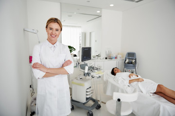 Obraz premium Examination is over. Portrait of beautician in white lab coat looking at camera and smiling. Brunette woman in bathrobe lying on daybed on blurred background