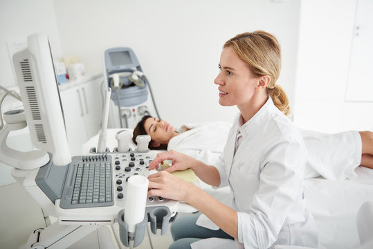 Side view portrait of smiling physician in white lab coat looking at monitor of ultrasound machine. Brunette woman with closed eyes lying on daybed on blurred background