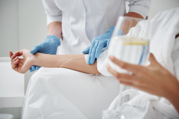 Close up of physician hand in sterile glove checking tube and needle for IV infusion on woman arm. Girl holding glass with lemon water - obrazy, fototapety, plakaty