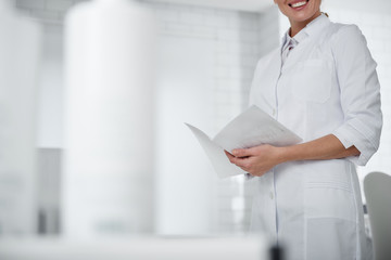 Cropped portrait of young cosmetologist in white lab coat holding brochure and smiling. Copy space...