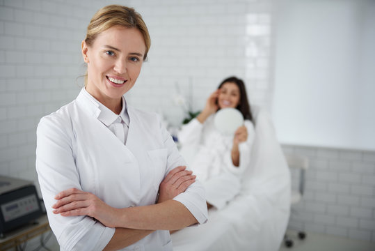 Waist up portrait of charming beautician in white lab coat crossing arms and looking at camera with smile. Young woman in bathrobe looking in the mirror on blurred background