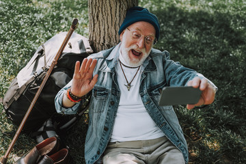 Waist up portrait of joyful hipster old male resting on grass near tree during skype connection