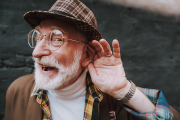 What. Close up portrait of stylish pensioner with playful glance holding palm near his ear and...