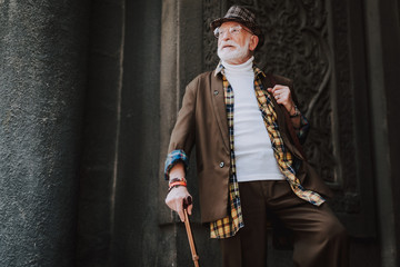 Waist up portrait of happy hipster senior manstanding on stairs of grand building while holding stick