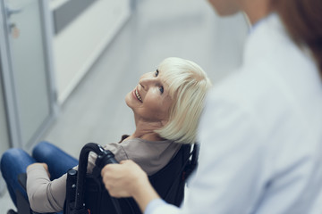 Top view with focus on delighted senior woman sitting in wheelchair. She is looking up at nurse...
