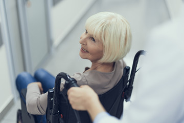 Top view with focus on joyful mature woman sitting in wheelchair. Medic is standing behind and assisting her by pushing vehicle