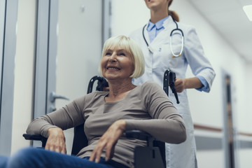 Smiling aged lady is sitting in pushchair while visiting hospital. Therapist is helping her by...