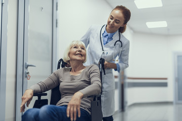 Smiling medical worker is propelling pushchair of happy lady while moving in clinic. They are looking at each other with joy while patient is sitting and professional is standing behind