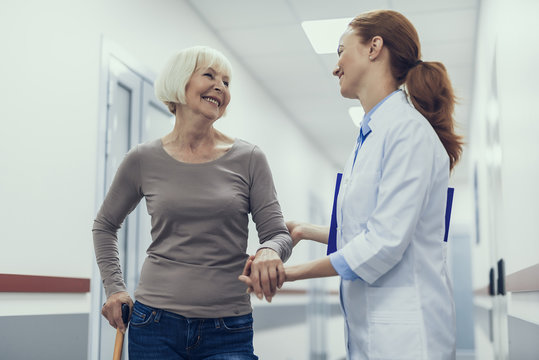 Low angle of cheerful mature lady and female therapist communicating in clinic. They are standing together and medical worker is supporting woman by hand as she is using cane for walking
