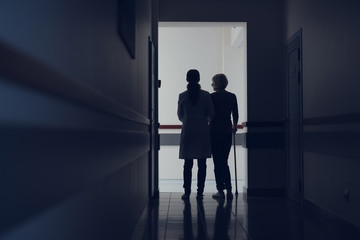 Lady with cane is leaning on medical worker hand. They are walking together through corridor. Copy...