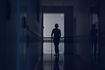 Woman is using crutch for moving in hospital. She is crossing dark hallway with focus on back. Copy space in left side