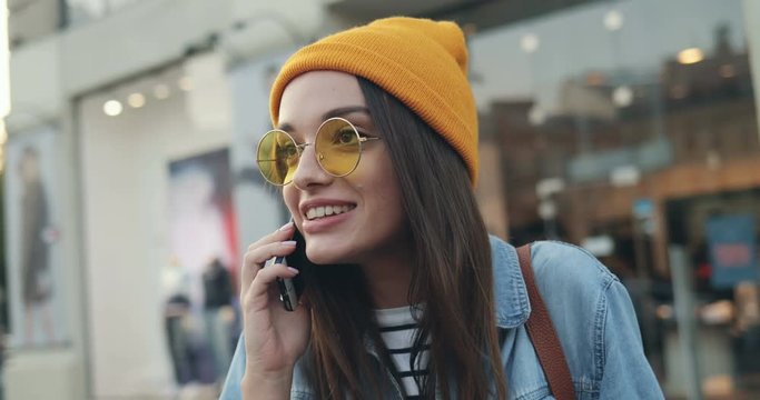 Close up of the young stylish girl in the sunglasses and hat talking on the phone with many emotions like explaining something on the street. Outdoors.