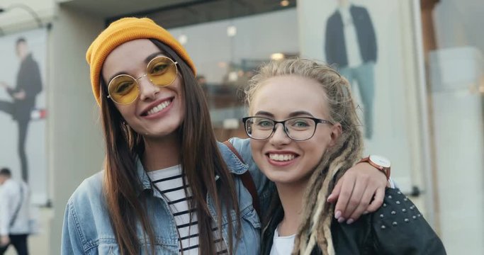 Portrait shot of the two stylish and pretty girls best friends in stylish looks posing to the camera cheerfully with smiles and hugging on the city street. Outside.