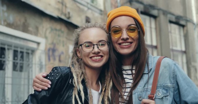 close up of the two attractive young Caucasian women best friends in hipster looks posing to the camera cheerfully with smiles and embracing on the city street. Outdoors Portrait..