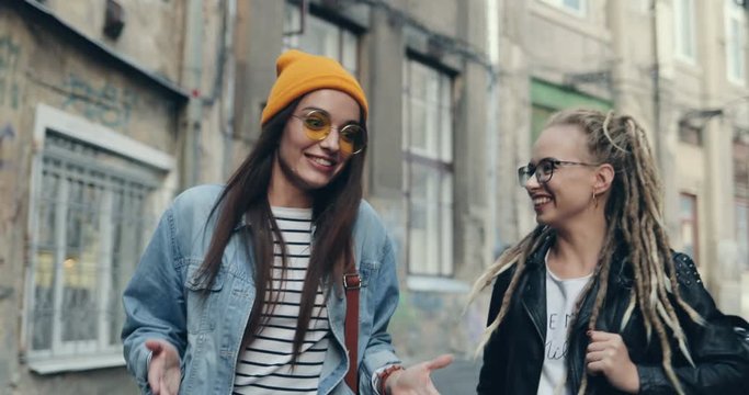 Two Caucasian cheerful hipster girls friends walking the street together happily and talking emotionally. Outside.