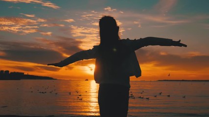 Silhouette of happy child girl stands on background of sunset sky and sea and throws his hands as if flies. Summer vacation, adoption and people concept. Happy little girl having fun over sunset.
