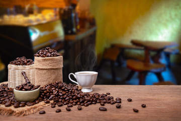 Naklejka na ściany i meble Hot cup of coffee and beans with burlap sack on the wooden table, with coffee shop background. copy space