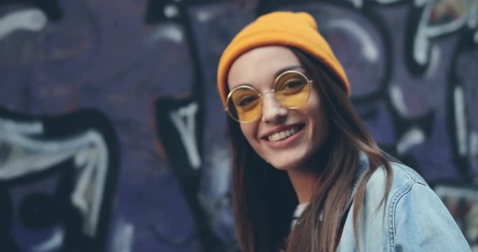 Back view of the close up of the hipster stylish young girl in hat and sunglasses turning her head smiling cheerfully to the camera on the graffity background. Close up. Portrait. Outside.