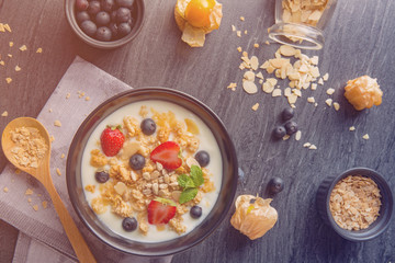 bowl of oat granola with yogurt, fresh blueberries, strawberries, blackberries and nuts with spoon on black wooden board for healthy breakfast, top view