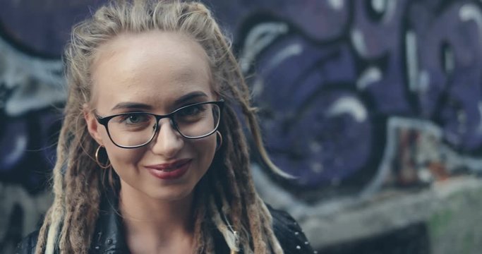 Close up of the camera zooming in the face of the pretty stylish Caucasian girl in glasses and with dreadlocks standing on the graffity wall background and smiling. Outdoors. Portrait shot.