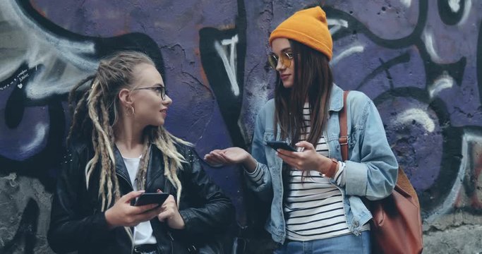 Two Caucasian stylish hipster female best friends standing on the graffity wall background, scrolling ot typing on their smartphones and talking. Outsside.