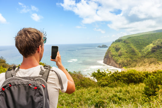 Travel tourist man in Hawaii beach USA vacation taking photo with mobile phone device of ocean landscape mountains background. Pololu Valley beach hike, Big Island, Hawaii.