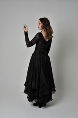 full length portrait of brunette girl wearing long black lace gown with corset.  standing pose with back to the camera, grey studio background.