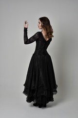 full length portrait of brunette girl wearing long black lace gown with corset.  standing pose with back to the camera, grey studio background.