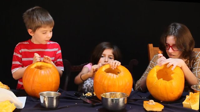 Three kids intently carving their Halloween pumpkins.  They are using small, saw like knives.  They are focused on their projects.