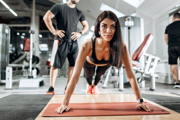 Naklejka na ściany i meble Beautiful athletic girl dressed in black sports top and tights is doing plank under the supervision of a coach in the gym