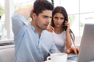 Displeased young loving couple sitting in cafe using laptop computer talking by phone.