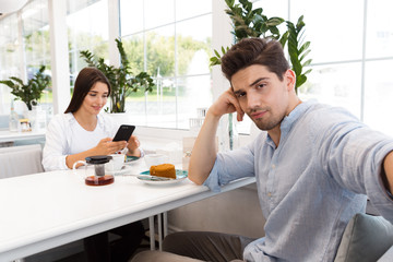 Fototapeta na wymiar Displeased man take selfie by camera while his girlfriend chatting by phone sitting in cafe.