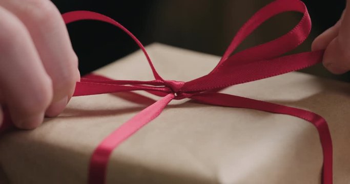 Slow motion macro shot of young female hands unties red ribbon bow on craft paper gift box