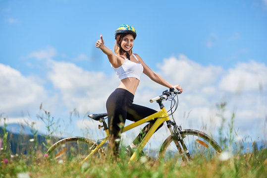 Smiling female cyclist riding on yellow bicycle in the mountains on summer day, wearing helmet, showing thumbs up sign against blue sky and clouds. Outdoor activity, lifestyle concept. Copy space