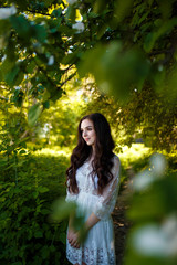 a young teenager girl with long dark hair in a white dress standing under a blooming apple tree