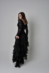 Fototapeta premium full length portrait of brunette girl wearing long black lace gown with corset. standing pose with back to the camera, grey studio background.