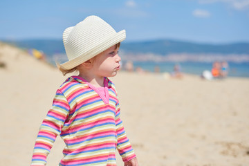 Little two year old girl with blue eyes in a bright wicker hat, standing on the beach near the sea