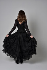 full length portrait of brunette girl wearing long black lace gown with corset.  standing pose with back to the camera, grey studio background.