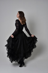 full length portrait of brunette girl wearing long black lace gown with corset.  standing pose with back to the camera, grey studio background.