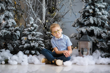Christmas portrait of happy child boy with big glasses siting on the floor indoor studio, snowy winter decorated tree on background. New Year Holidays