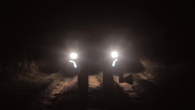 Silhouette of criminal man with axe standing at night road in light of headlight moving car, concept of robbery on road